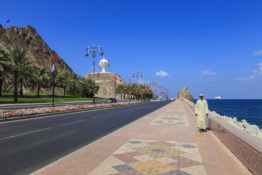 Man wearing dishdasha walks along Mutrah Corniche with national flags, flower beds and Giant Incense Burner, Muscat, Oman